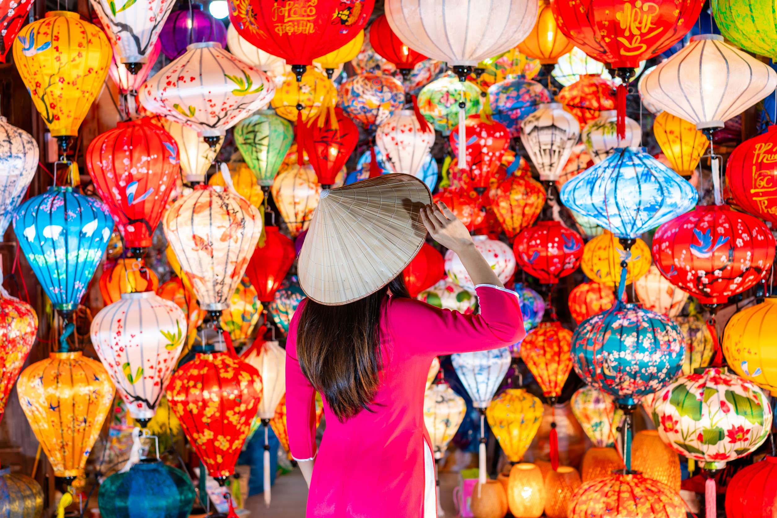 Young female tourist in Vietnamese traditional dress looking at paper ornamental lanterns in Hoi An